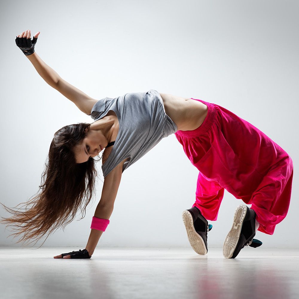 Woman in pink pants, gray top, dancing with arm raised, back arched, indoors.