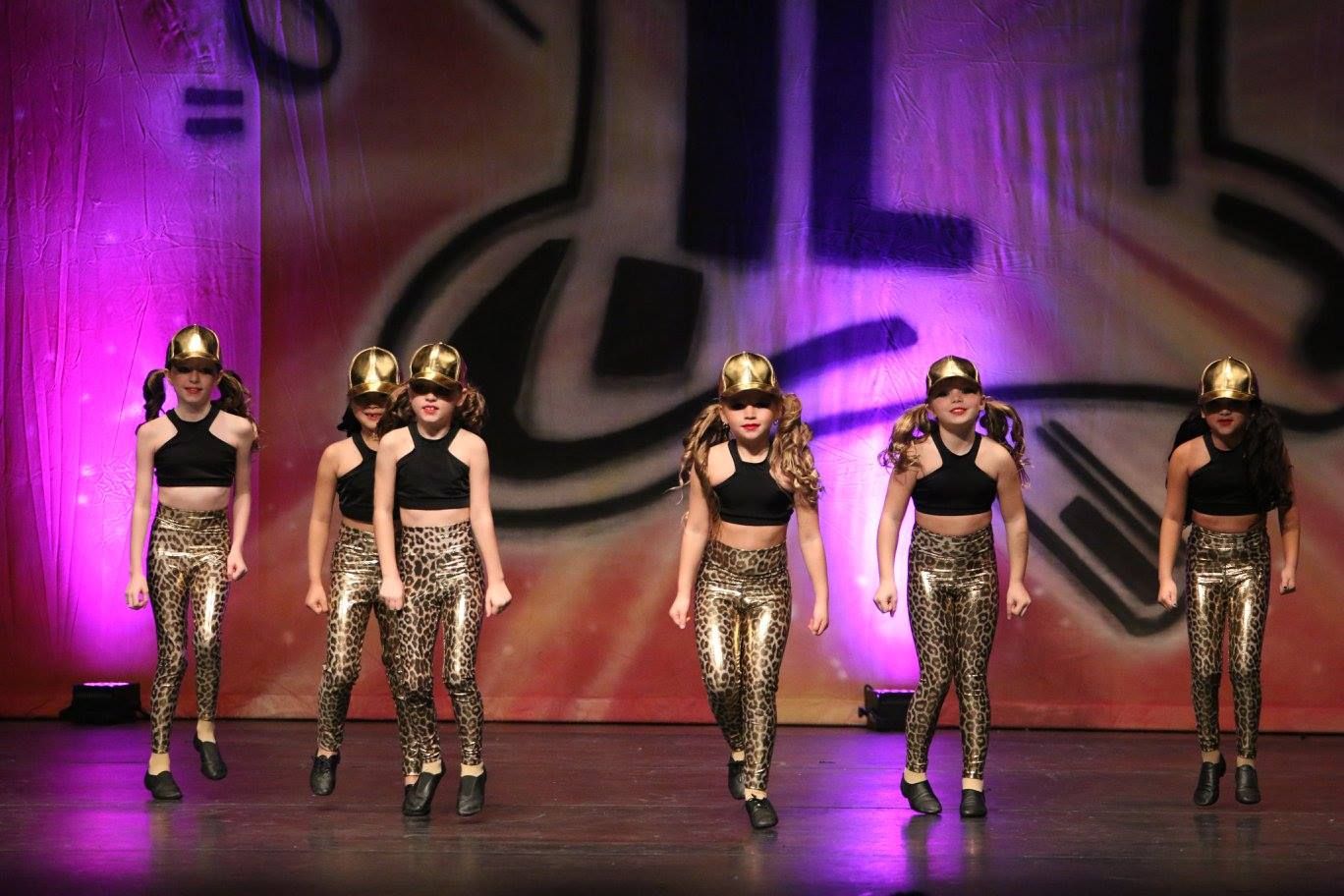 Group of children in dance costumes making heart shapes. They're on a stage, some with props.