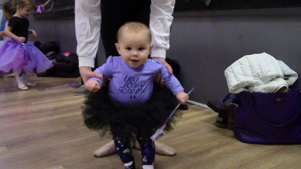 Young child in purple tutu and ballet slippers stands by dance studio mirror, holding a wand.