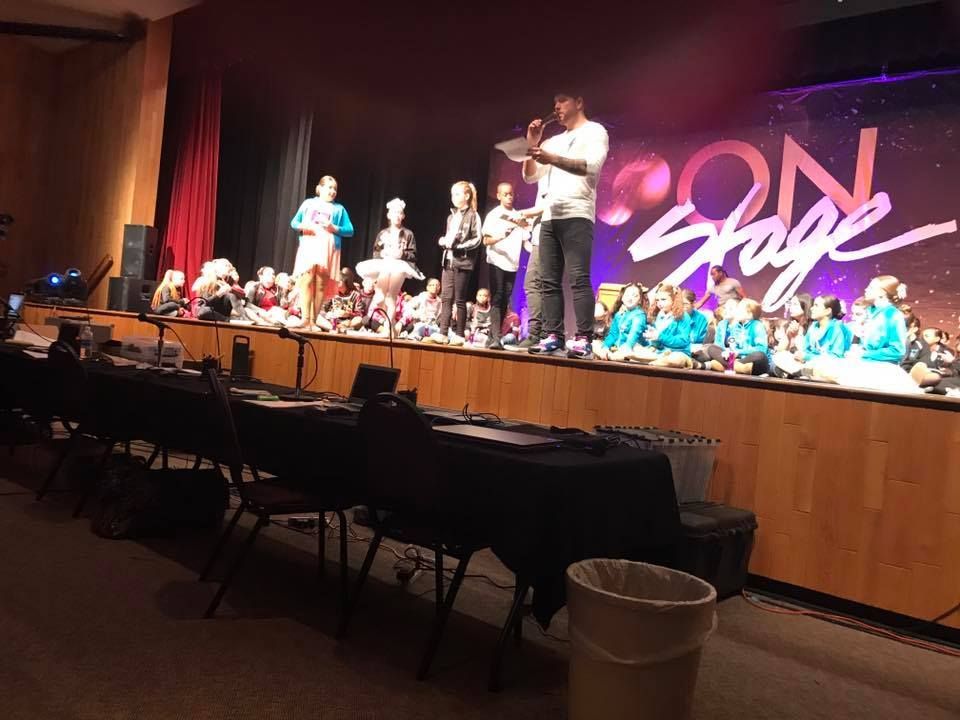 Group of children in dance costumes making heart shapes. They're on a stage, some with props.