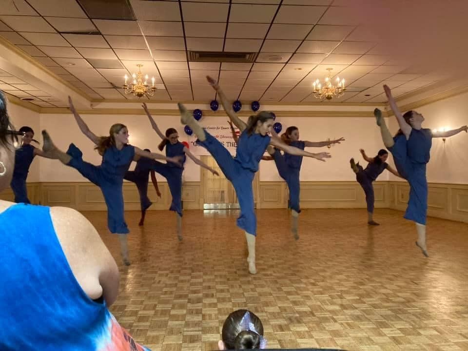 Group of children in dance costumes making heart shapes. They're on a stage, some with props.