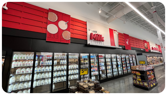 Grocery store dairy aisle with rows of refrigerated milk and dairy products, red sale signs overhead.