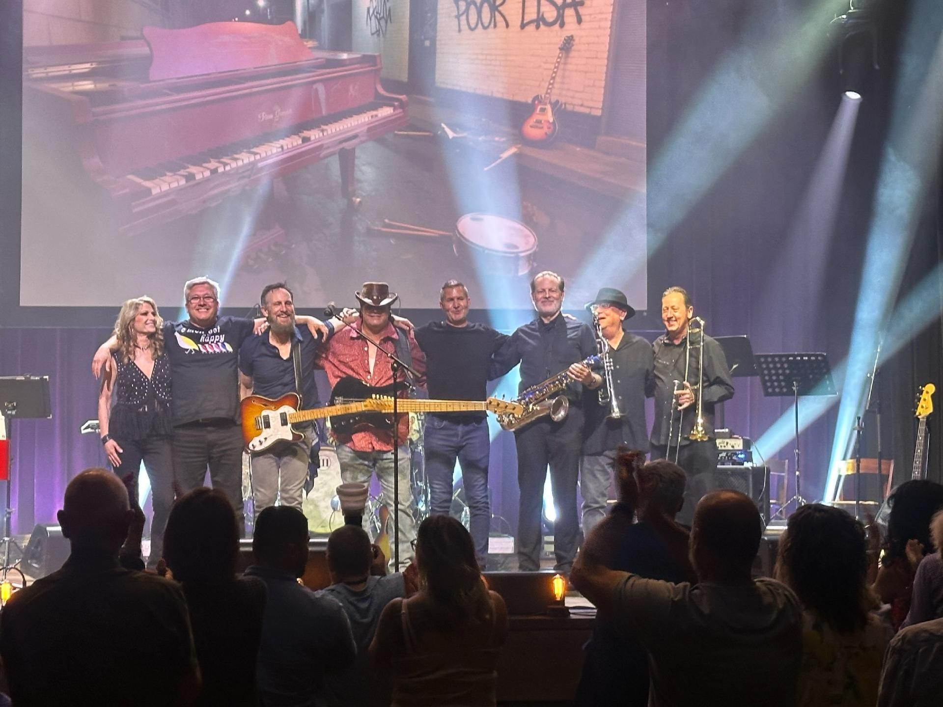 A band of eight musicians stands on a stage in front of an audience. A screen behind them displays a picture of a piano and the words A band of eight musicians stands on a stage in front of an audience. A screen behind them displays a picture of a piano and the words
