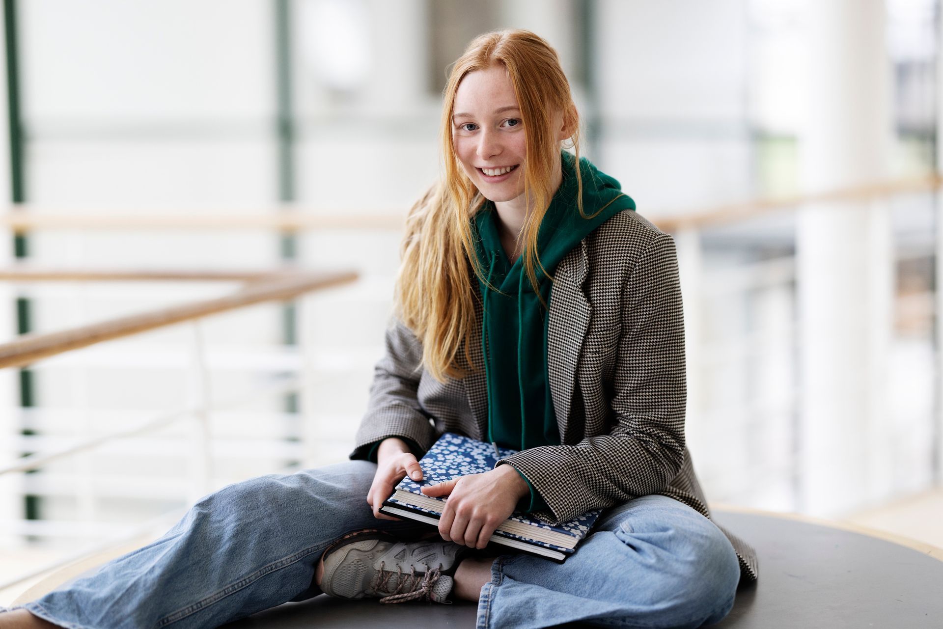 Woman with red hair, sitting, smiling, holding a small keyboard. Wearing a blazer, green hoodie, jeans. Bright, modern interior.