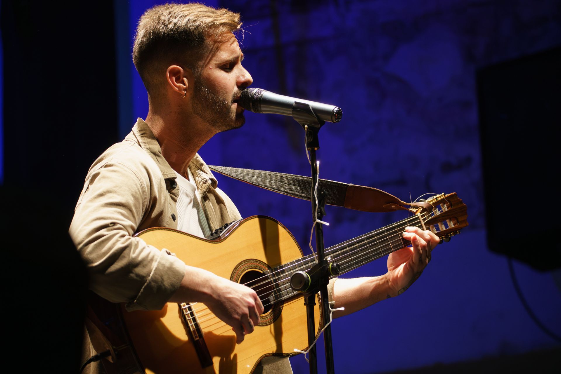 Man singing and playing acoustic guitar on stage, lit with blue light.