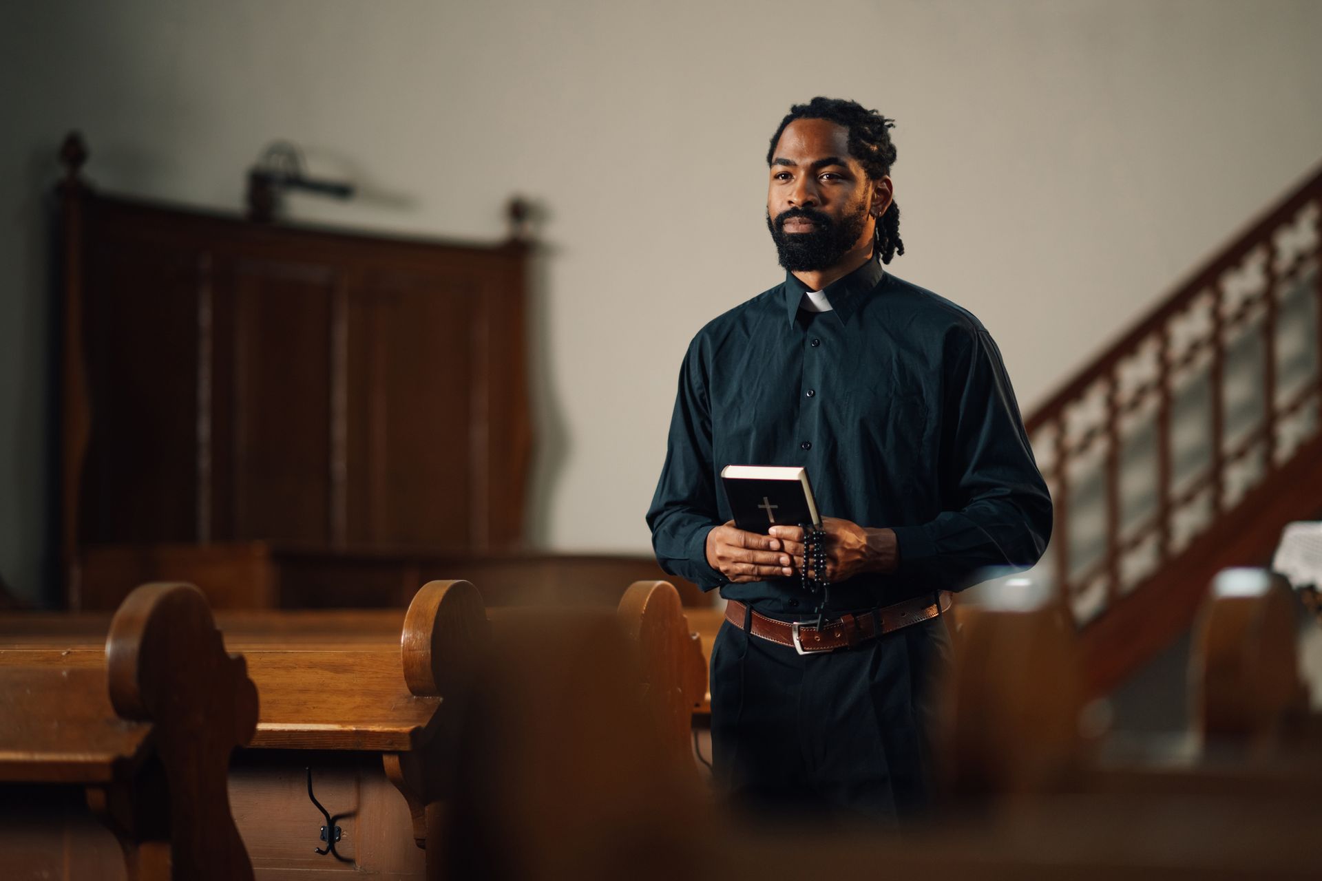 Man in clerical collar holds a book, standing in a church before pews.