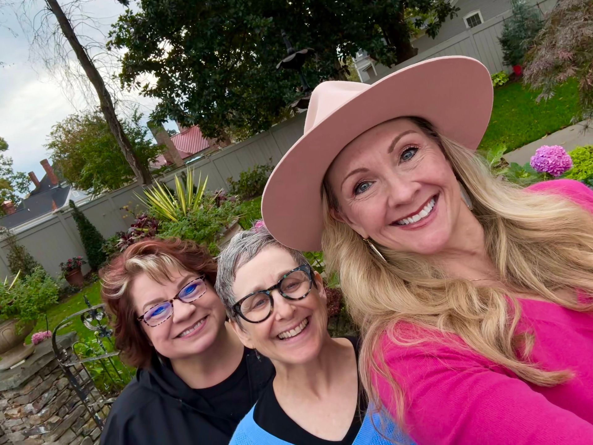 Three women are posing for a selfie in a garden . one of the women is wearing a pink hat. Three women are posing for a selfie in a garden . one of the women is wearing a pink hat.