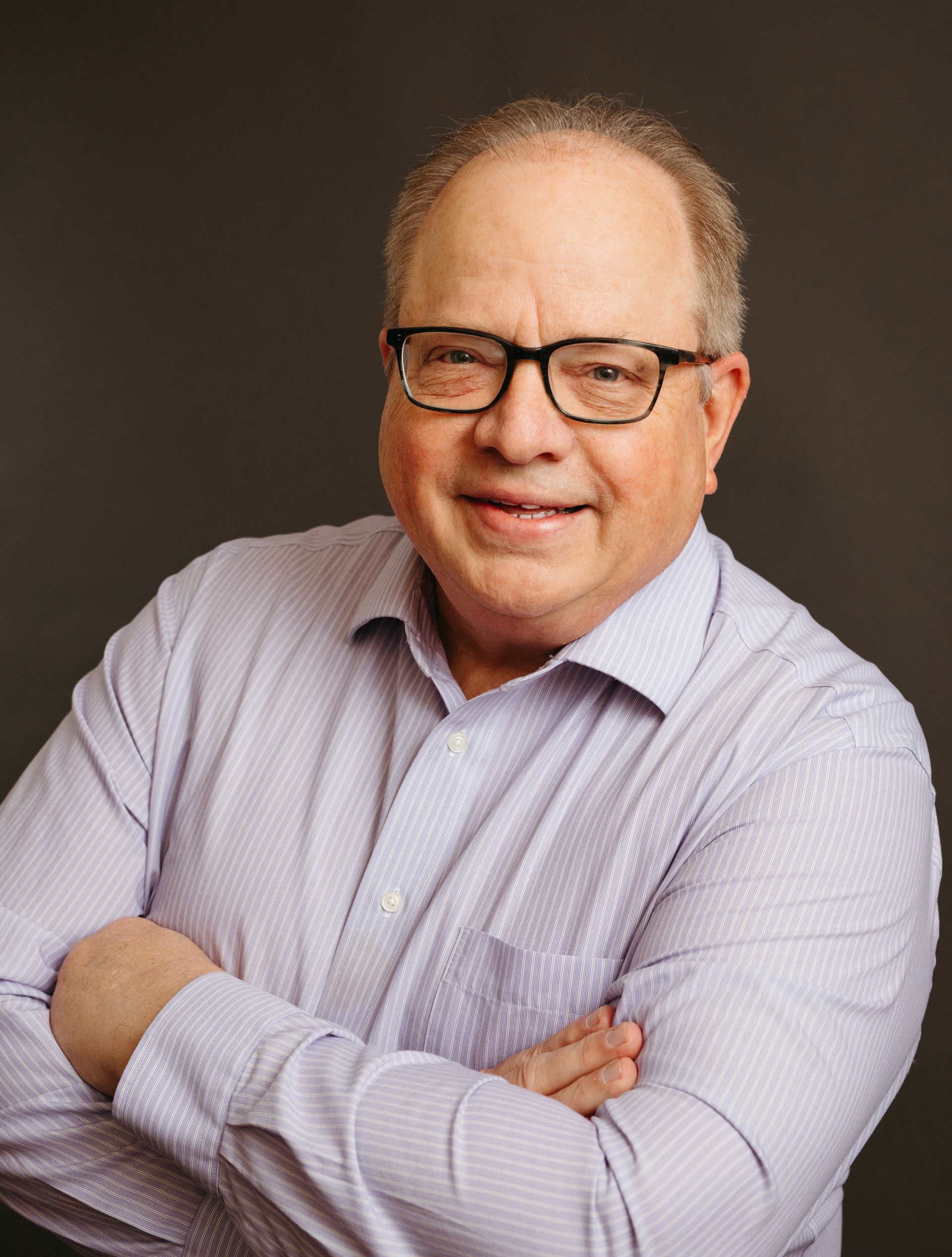 A man wearing glasses and a purple shirt is standing with his arms crossed. A man wearing glasses and a purple shirt is standing with his arms crossed.