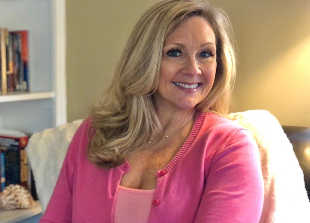 Woman with blonde hair smiles, wearing pink cardigan, sitting in a chair near a bookshelf. Woman with blonde hair smiles, wearing pink cardigan, sitting in a chair near a bookshelf.