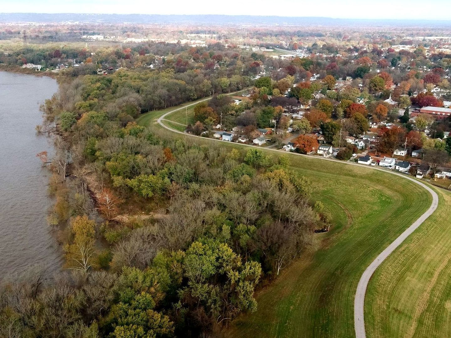 Lush shoreline and green levee curving along a river, with houses and fall foliage in the background.