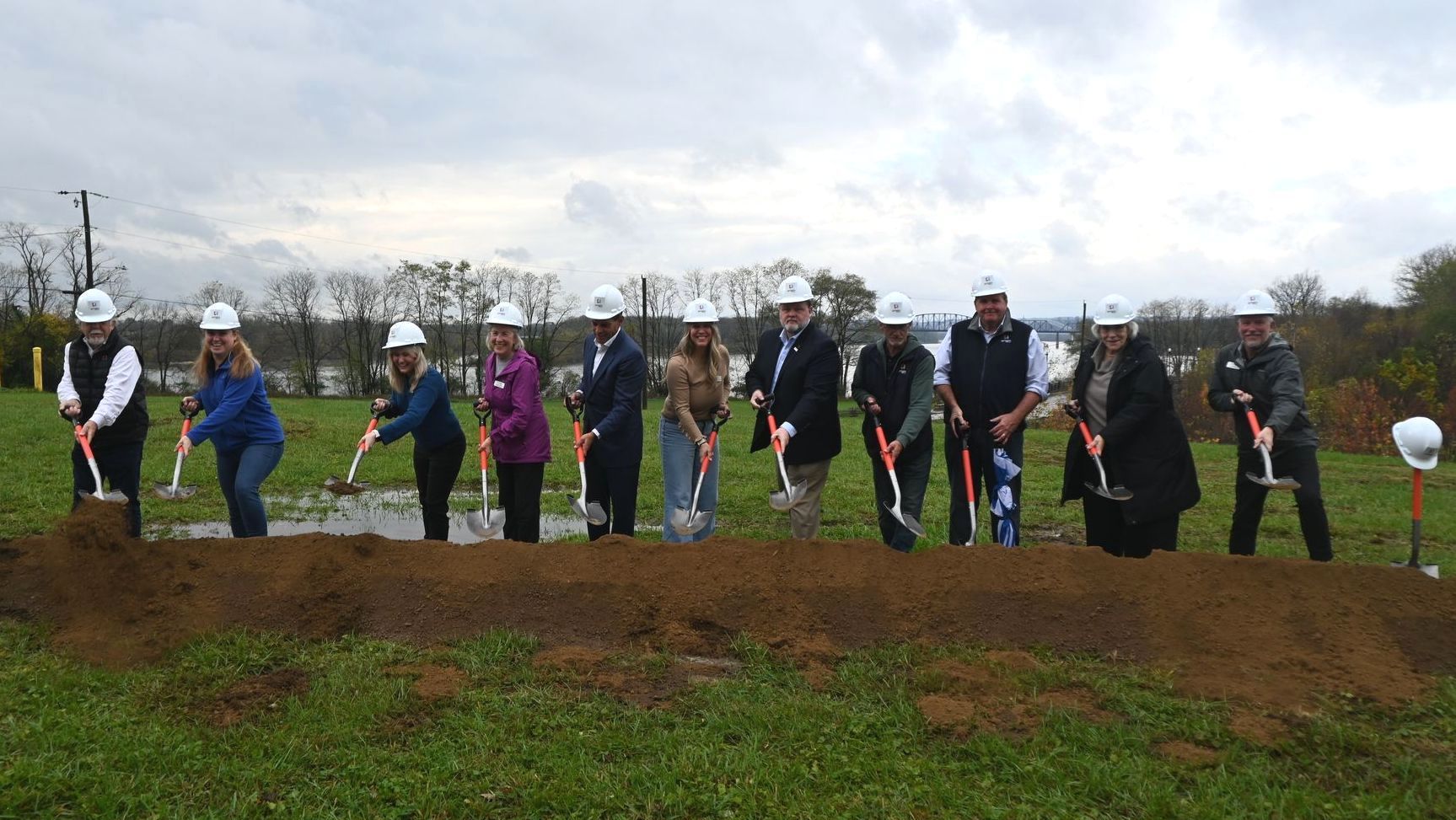 Groundbreaking of The Vista at Origin Park. River Heritage Conservancy staff members in hard hats and formal wear break ground with shovels on a grassy field under an overcast sky.
