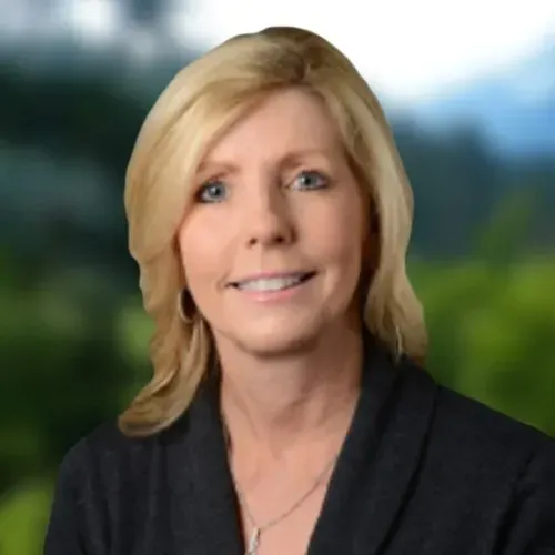 Dana Huber in a dark blazer smiles against a blurred outdoor backdrop.