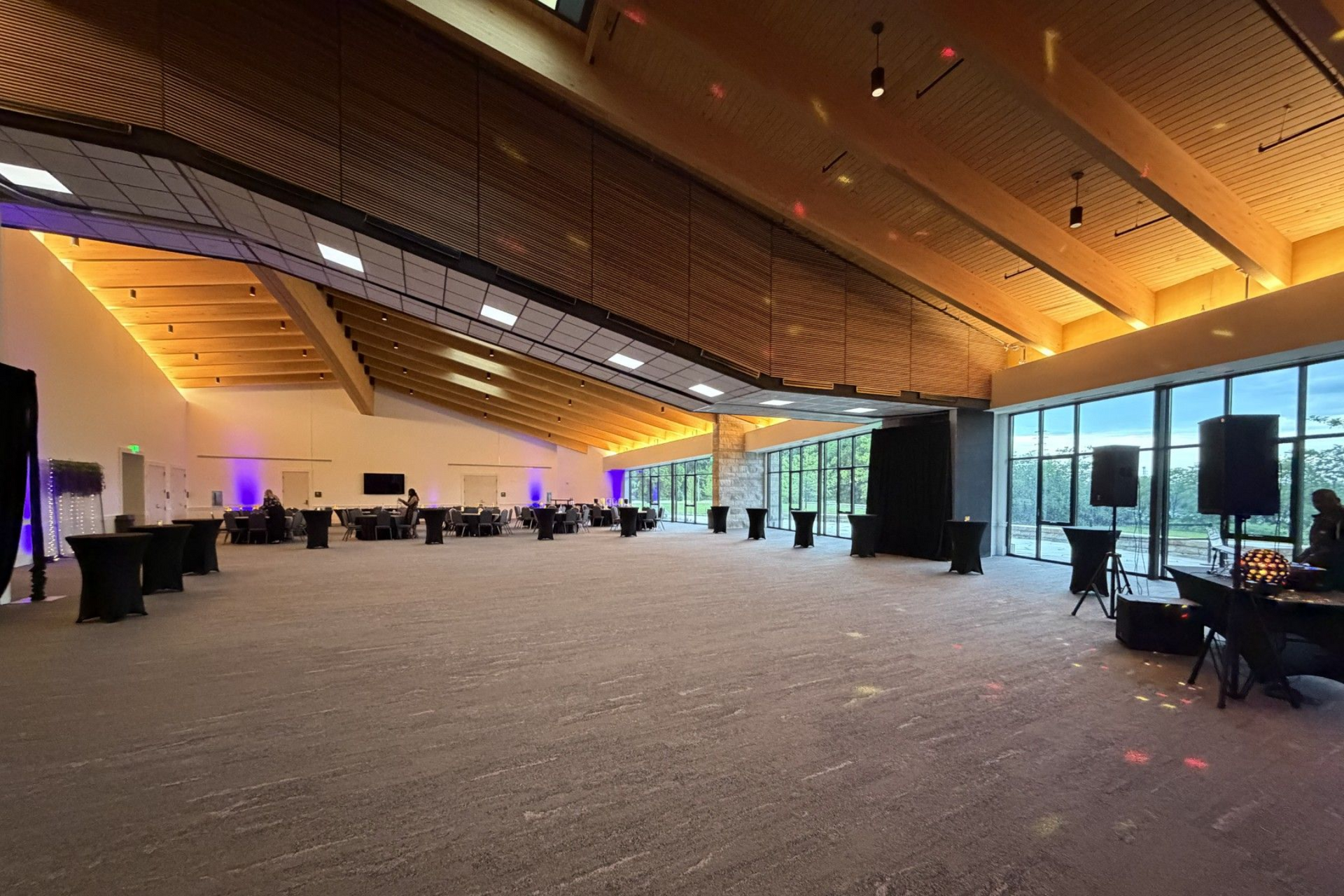 Conference room with people seated at a long table. One person stands, facing a screen. Windows and wood ceiling.