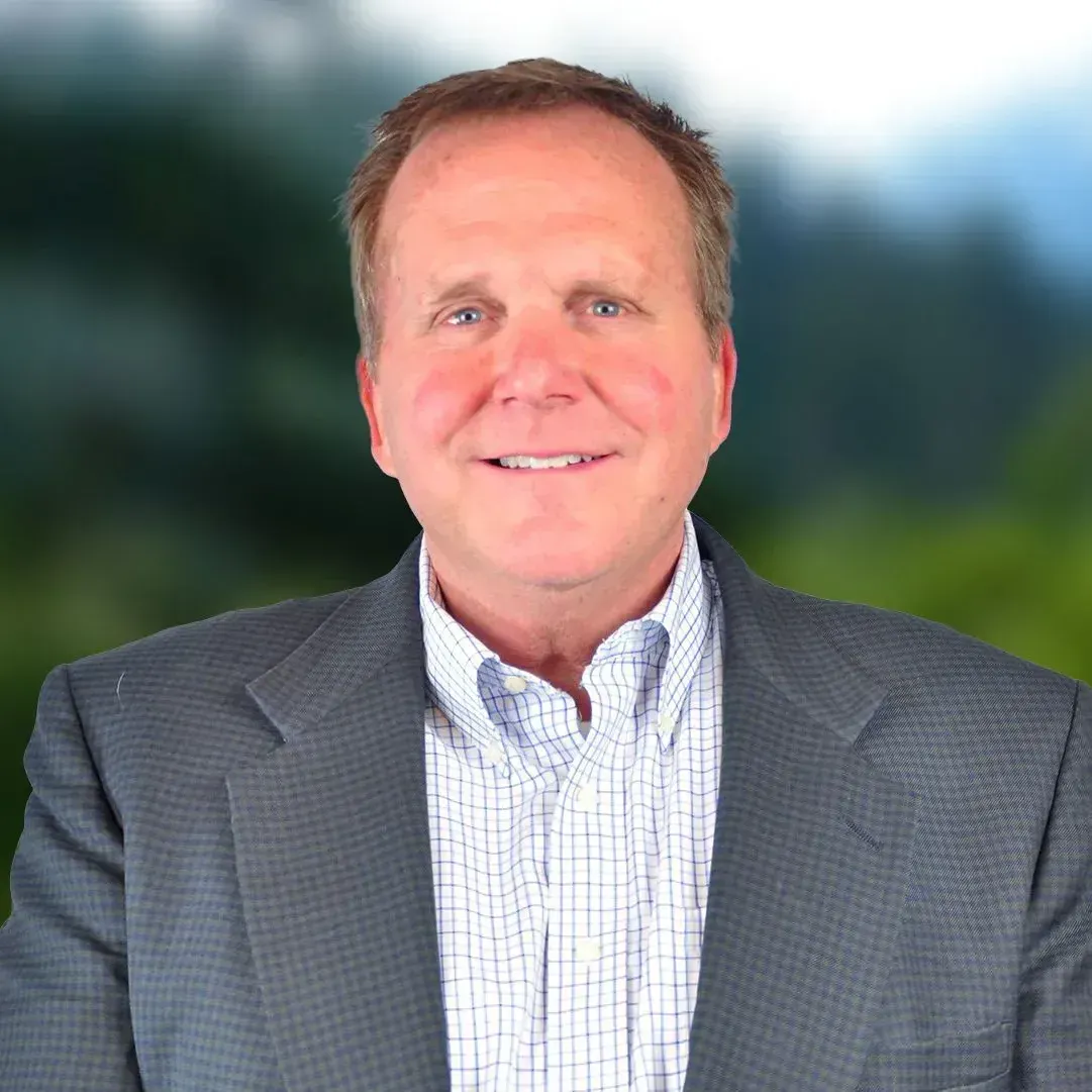 Kevin Beck in gray blazer and blue shirt smiles, with a blurred nature background.