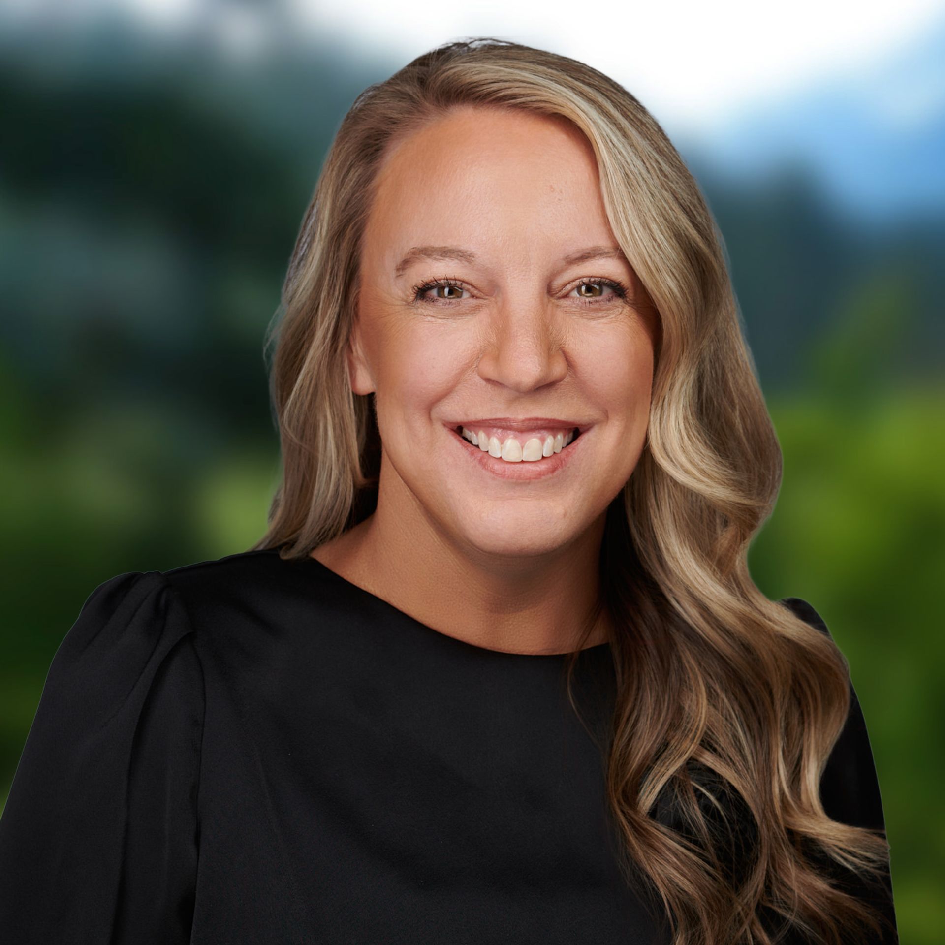 Shannon smiling woman with blonde hair in black dress, blurred green nature background.