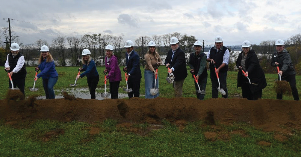 River Heritage staff members with shovels in a row, wearing hard hats, breaking ground on a cloudy day. Green grass, brown dirt.