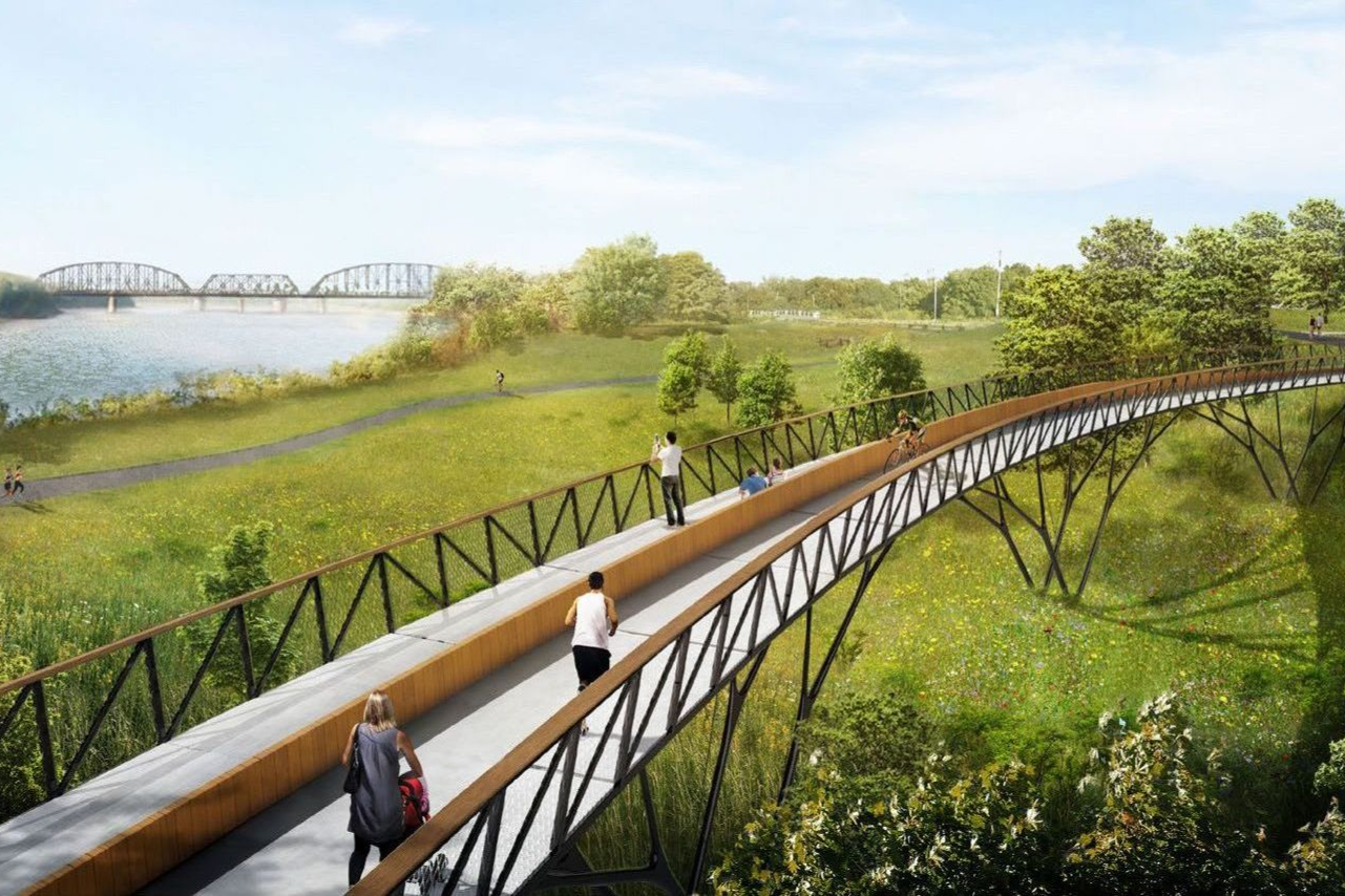 A walking bridge over a green landscape with people on it, next to a lake with a bridge in the background.