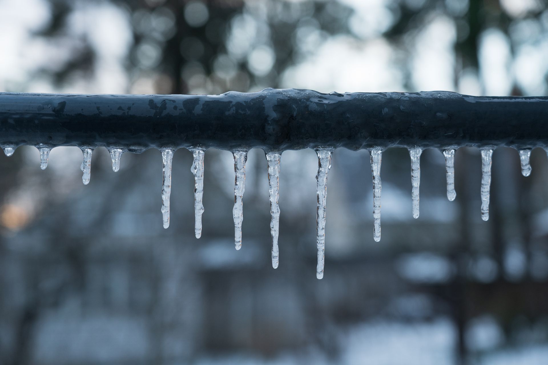 Icicles hanging from a dark pipe, blurry background of trees and buildings.