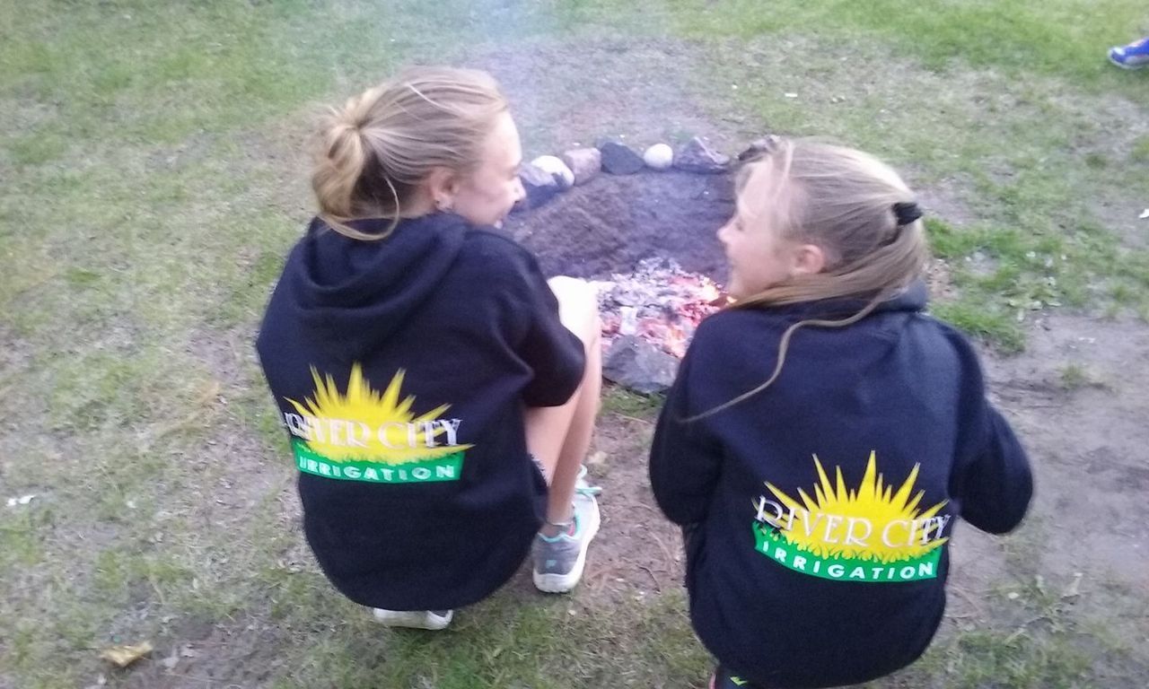 Two Girls Are Sitting in Front of A Fire Pit Wearing Sweatshirts that Say Sunny Federation | Onalaska, WI | River City Irrigation