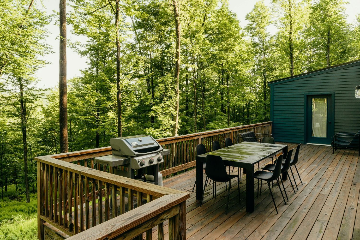 Wooden deck with grill and table set for dining, surrounded by lush green trees.