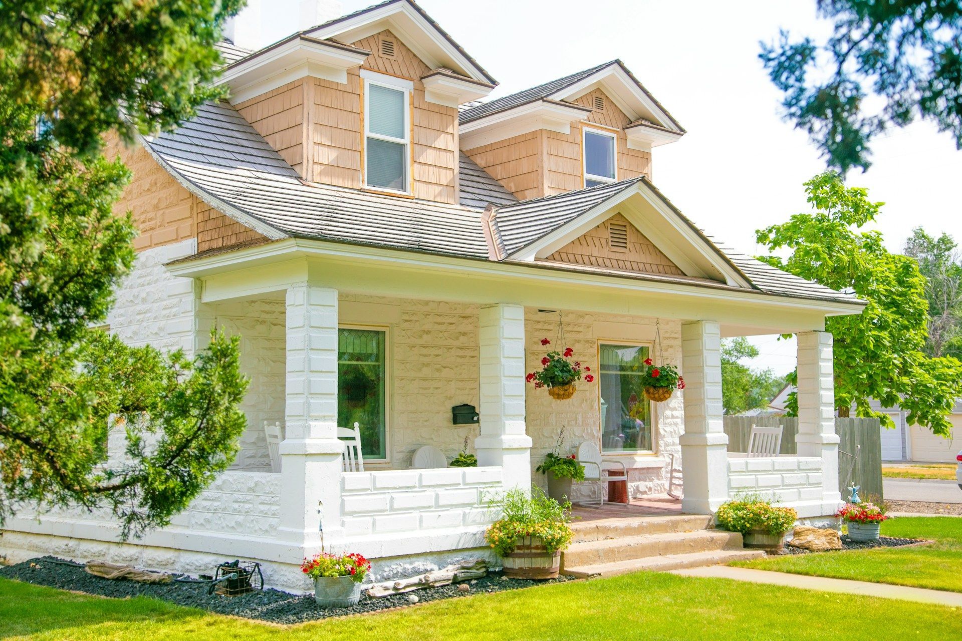 Two-story house with a white porch and light brown upper story, with green foliage 