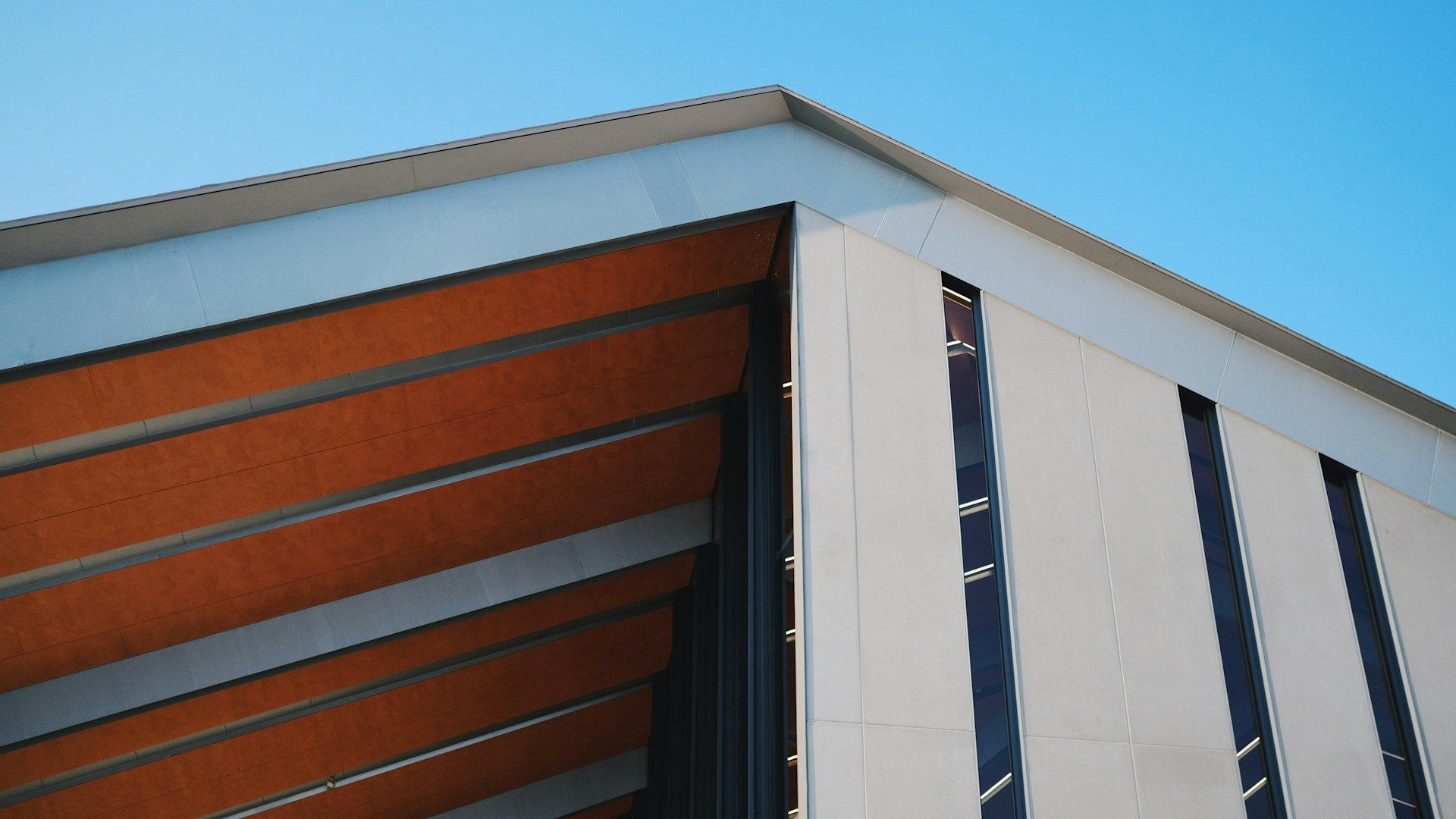 Corner of a modern building with long vertical windows and a contrasting wooden under-roof against a blue sky.
