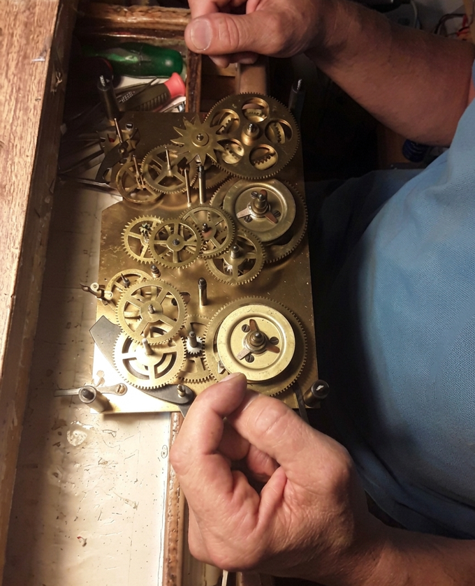 Mani che tengono gli ingranaggi di un orologio all'interno di una cornice di legno; gli ingranaggi sono color ottone e lo sfondo è una camicia blu.