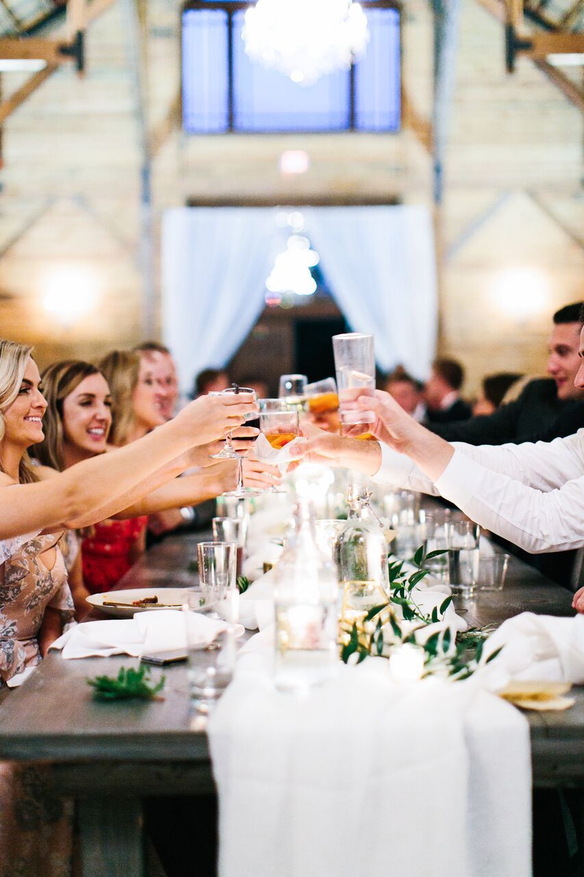 People seated at a long, rustic banquet table toast with glasses of drinks in a brightly lit, elegant indoor venue.