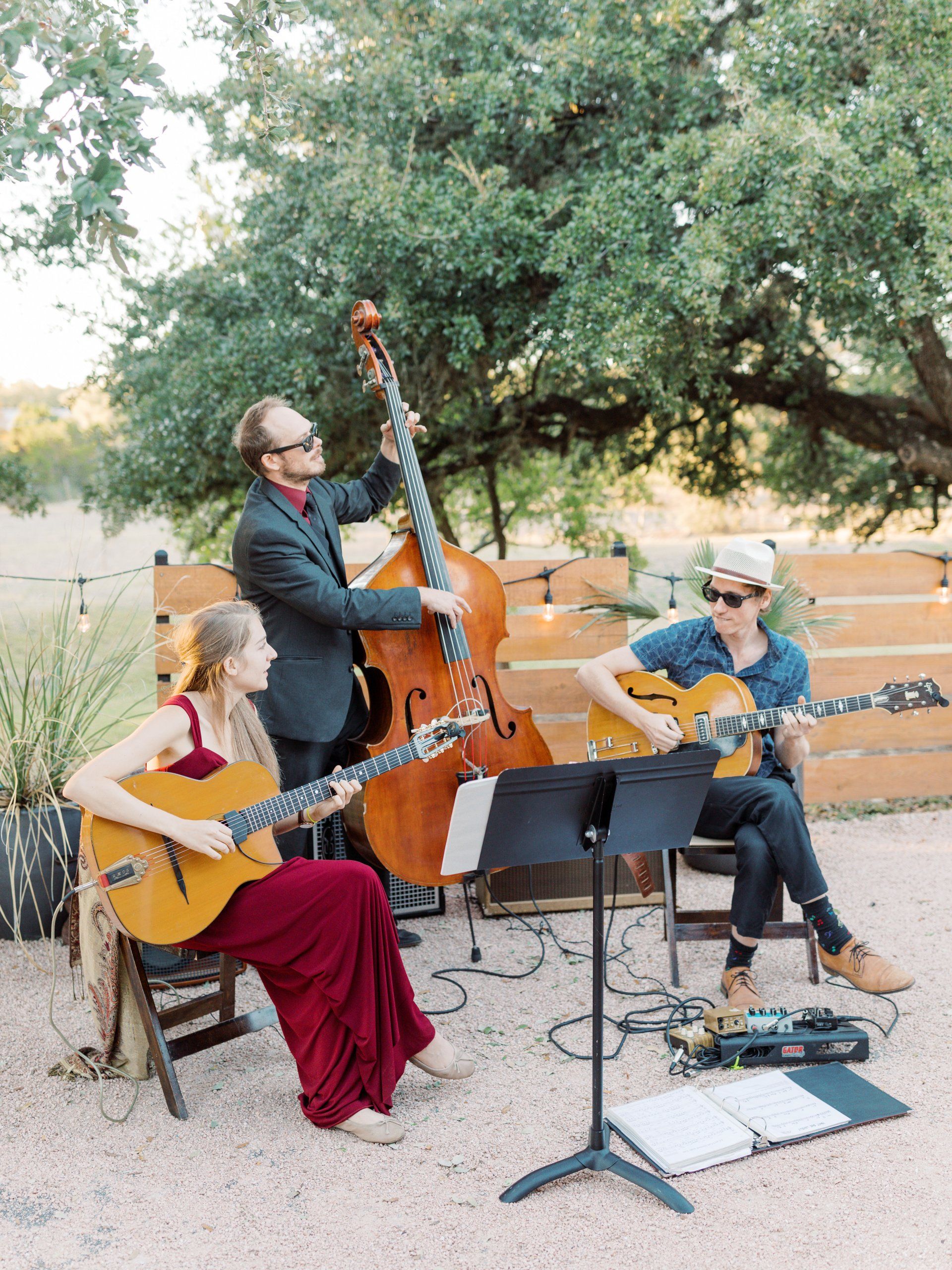 A trio plays acoustic guitar and upright bass outdoors in front of a wooden fence and large tree.