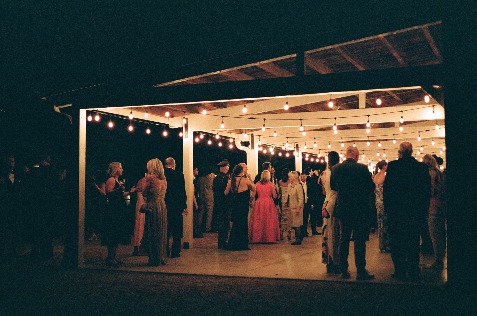 People gather under a roofed, open-air pavilion lit by hanging string lights at night.