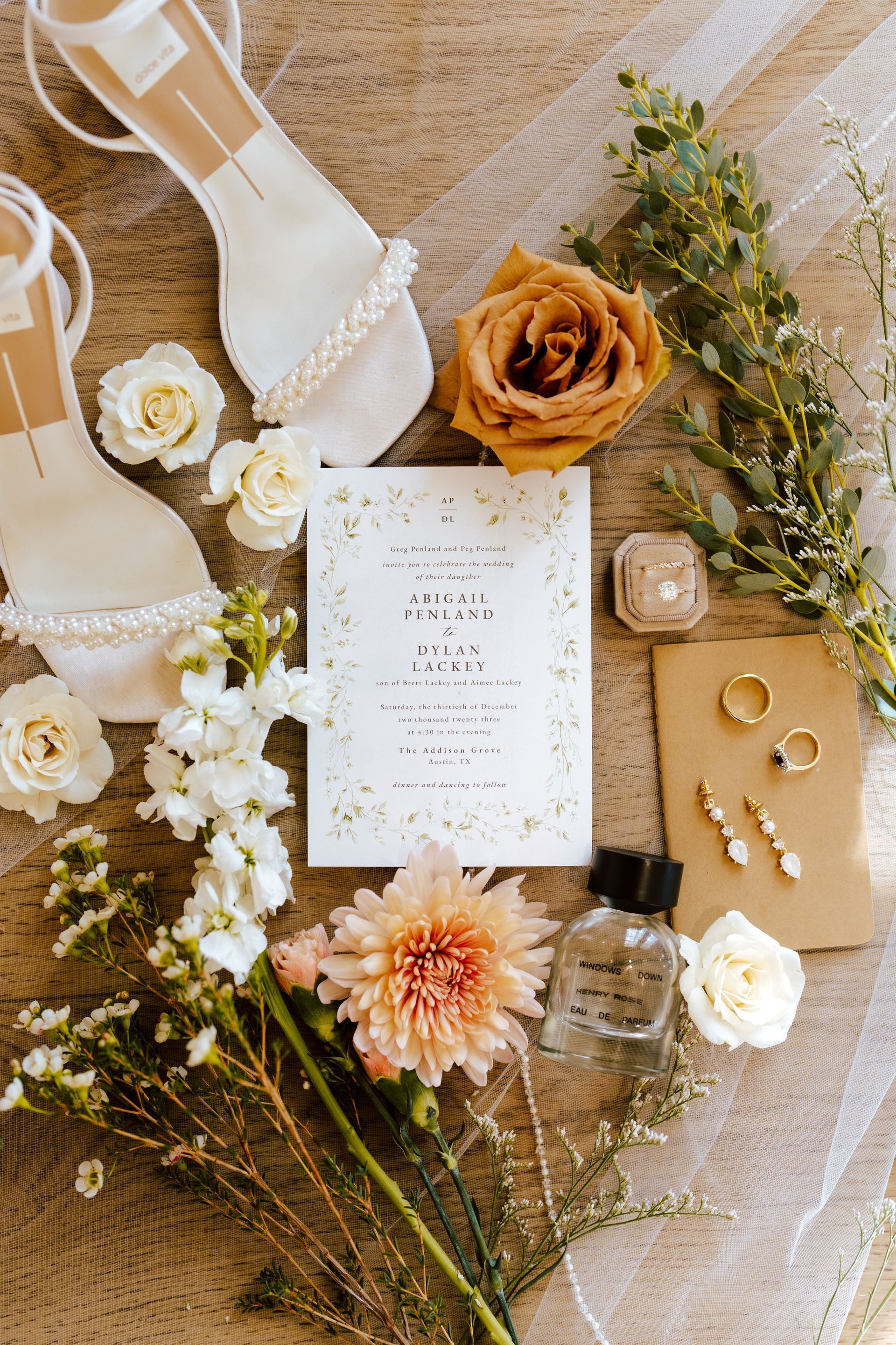 A flat-lay of wedding stationery, white heels, rings, earrings, a perfume bottle, and floral arrangements on linen.