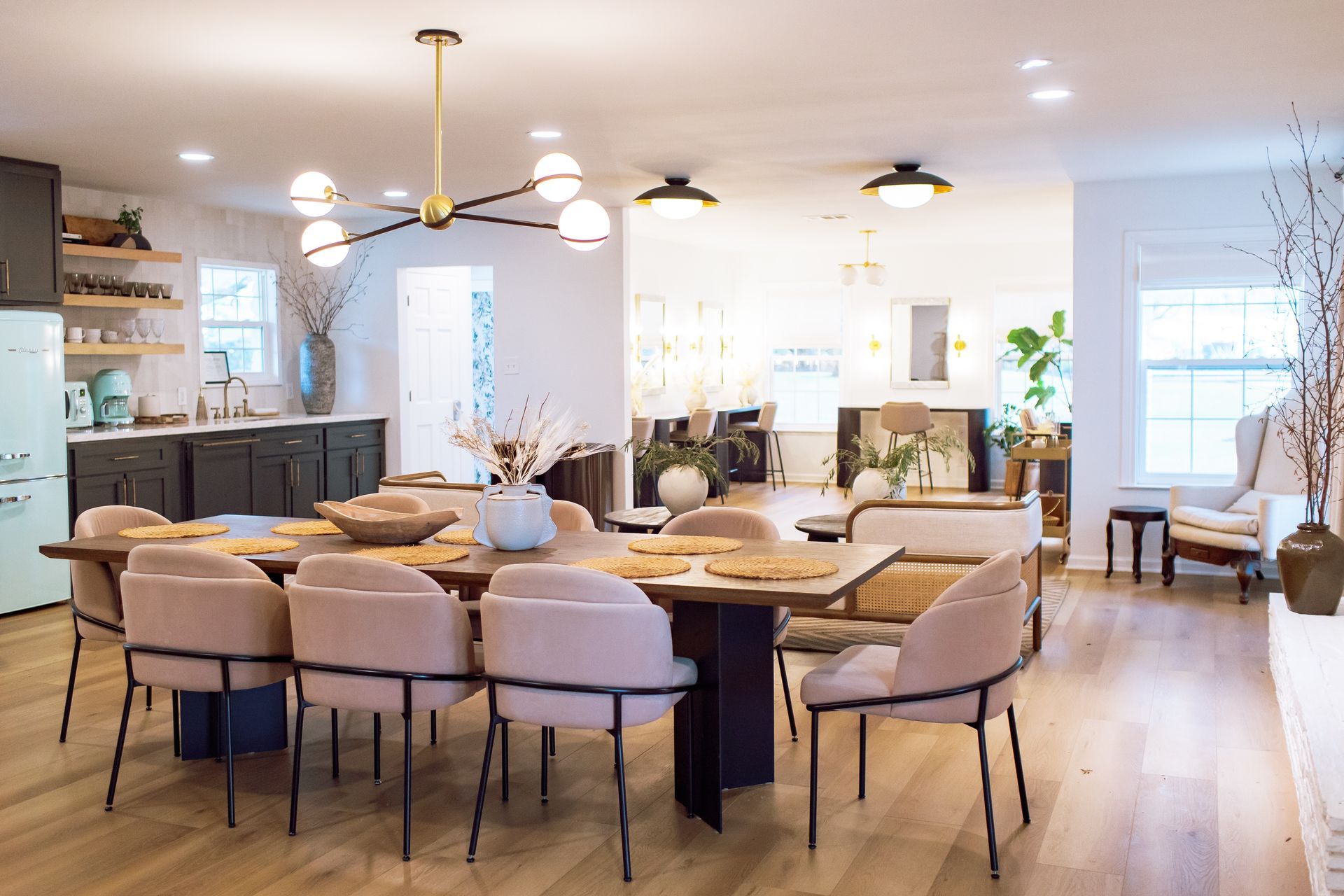 A bright, open dining area with a long wood table surrounded by beige chairs, under a modern chandelier, near a kitchen.