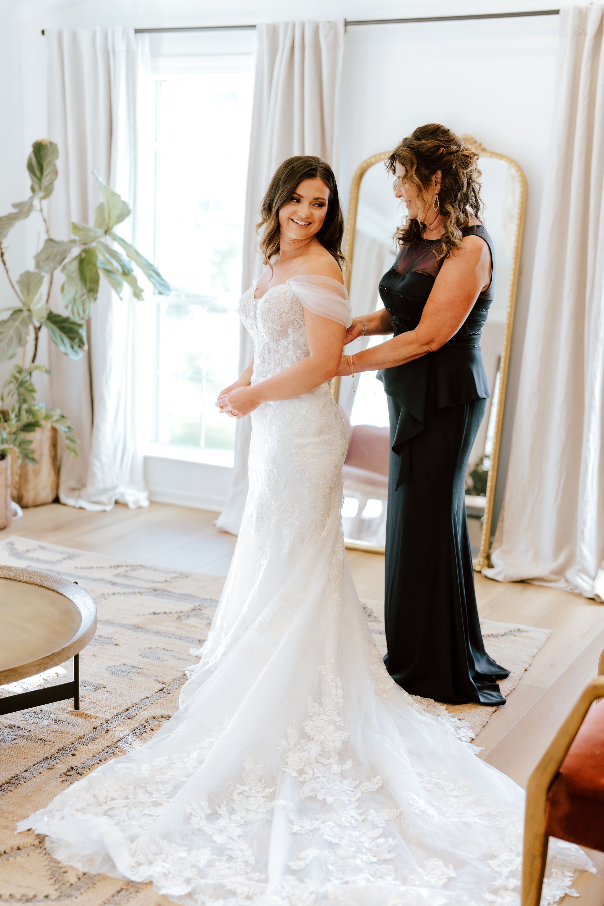 A person in a white wedding gown smiles as another person in a black floor-length dress adjusts the back of the gown.