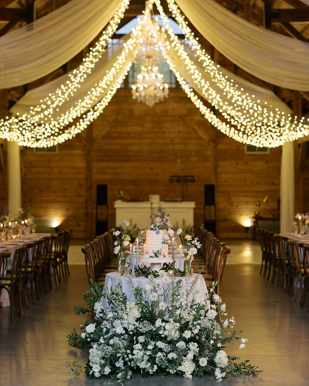 A wedding reception hall with draped fabric and fairy lights overhead, featuring a central floral-adorned cake table.