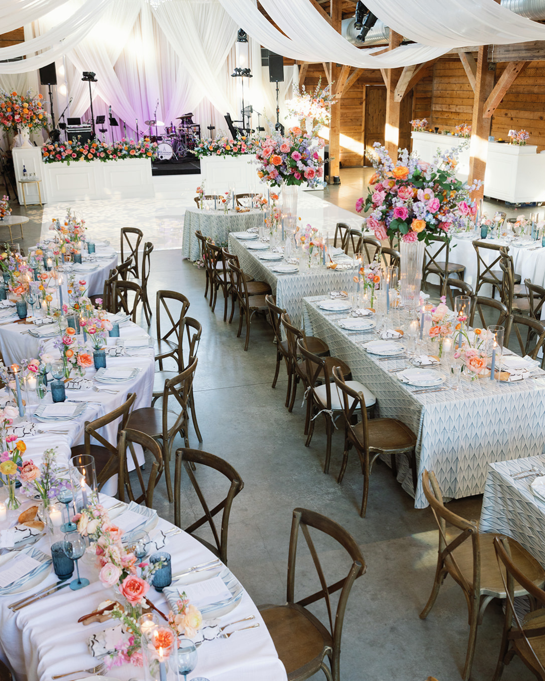 Elegant wedding reception with long floral tables, gold chairs, and draped white ceiling in a rustic barn.