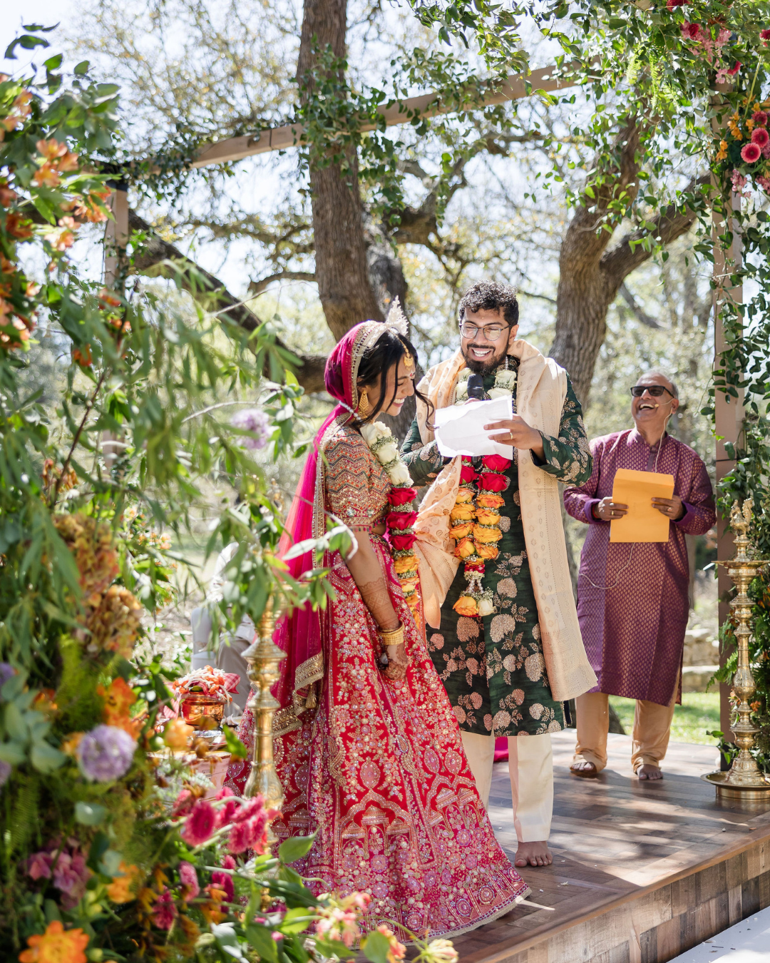 Two people in floral wedding attire stand on a decorated outdoor stage, reading from papers during a ceremony.