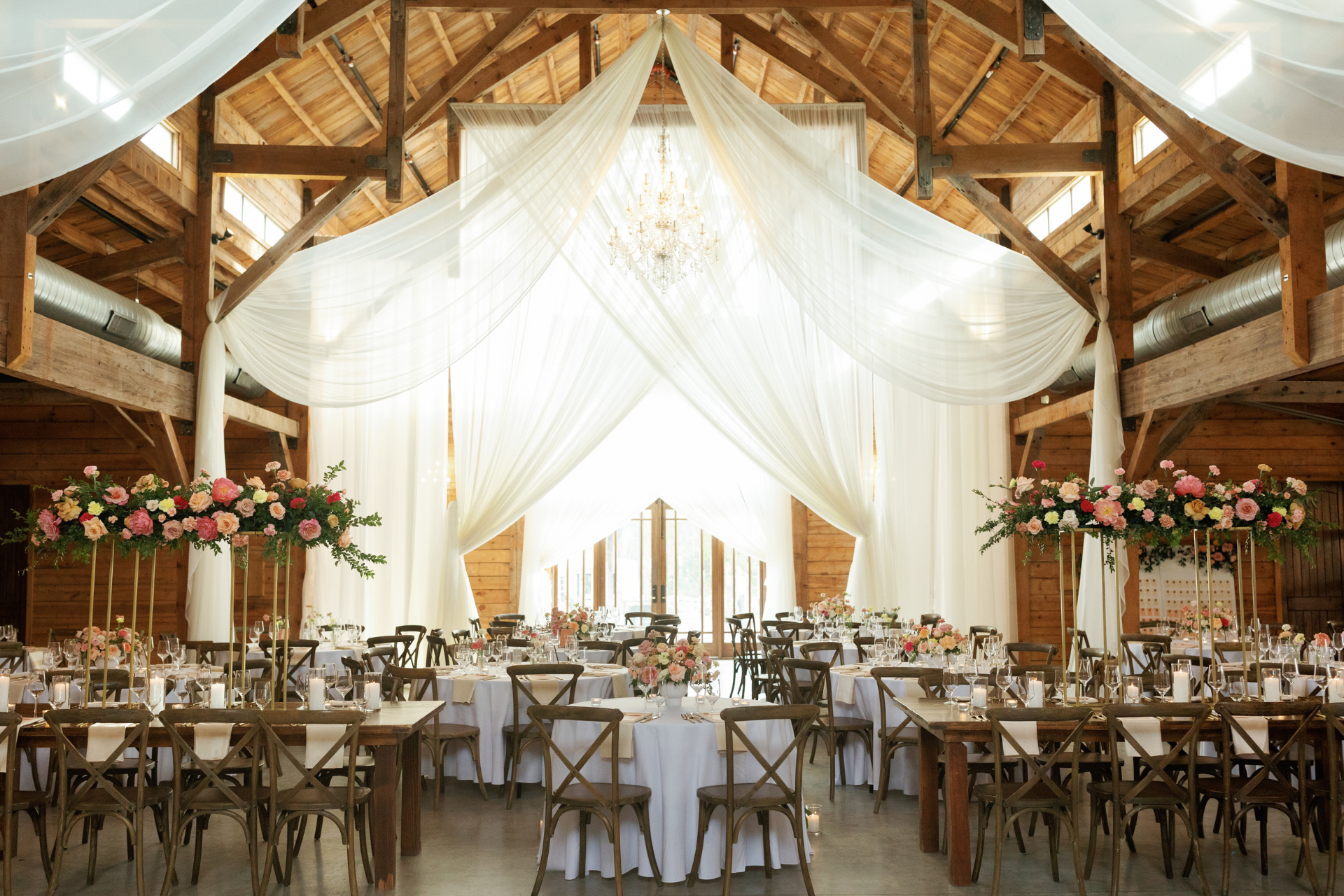 Rustic barn wedding reception featuring wooden beams, white draped fabric, chandeliers, and round tables with floral center.