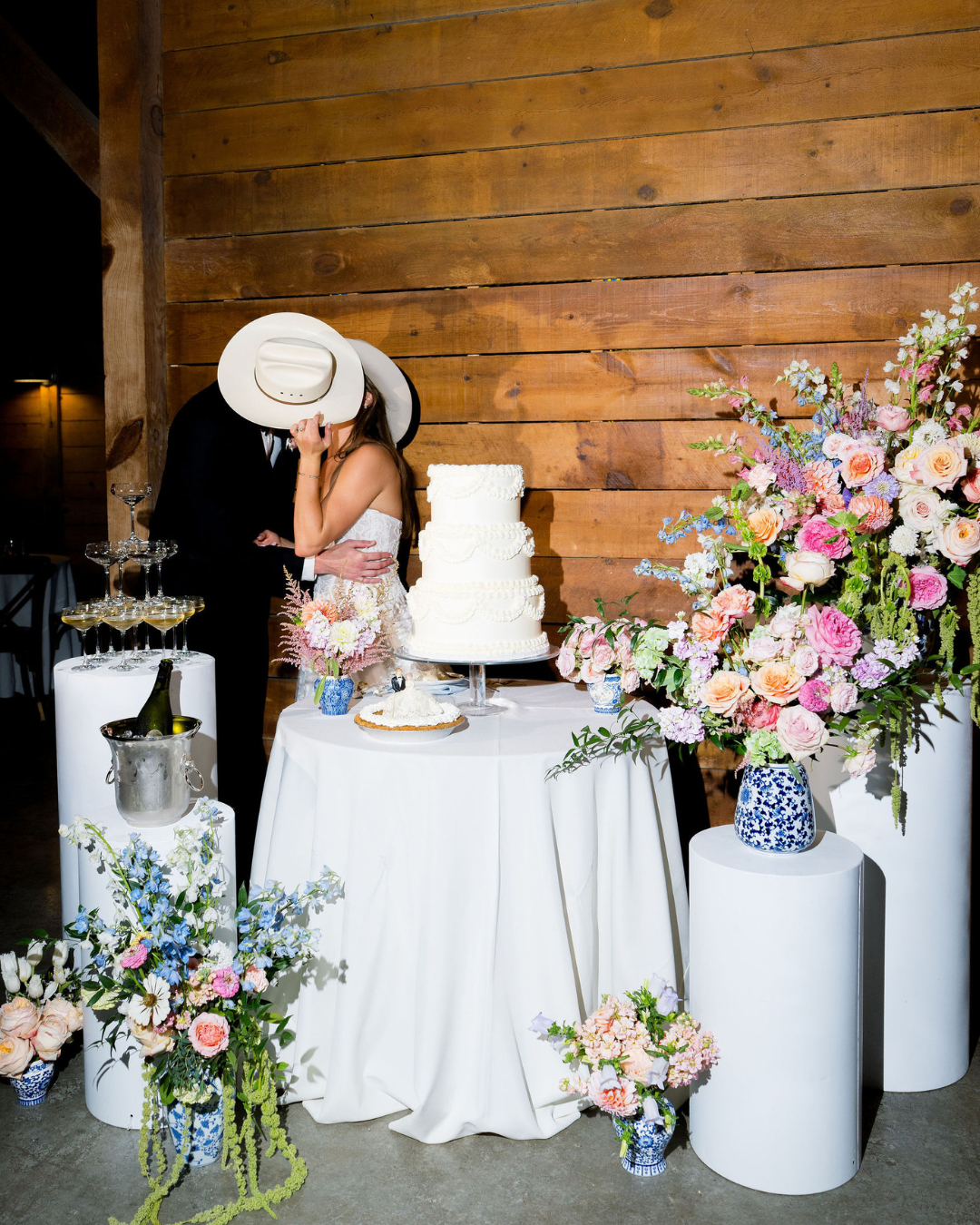 Wedding dessert table with white cake and pastel floral arrangements against a wood wall