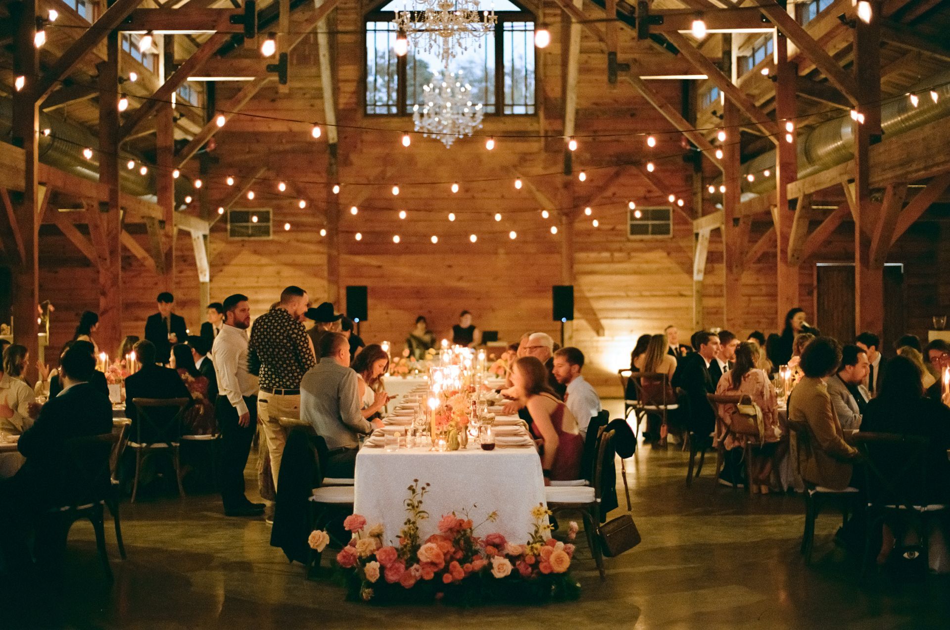 A rustic wedding reception inside a wood-paneled barn with string lights, a long white-clothed table, and guests seated.