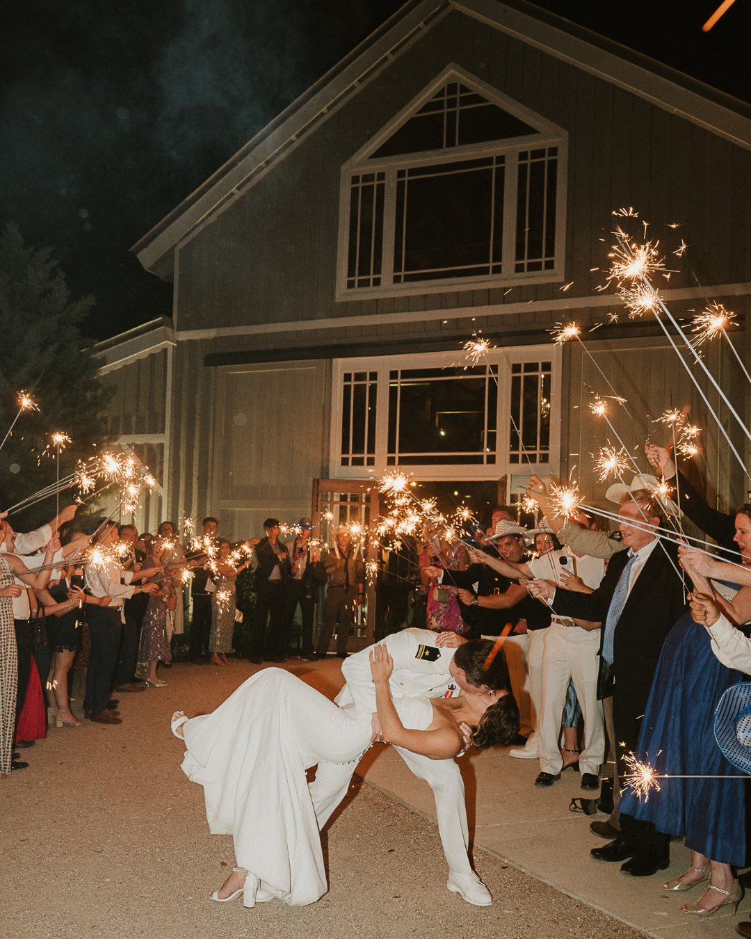 A couple in wedding attire shares a dip and kiss in front of guests holding lit sparklers outside a rustic barn at night.