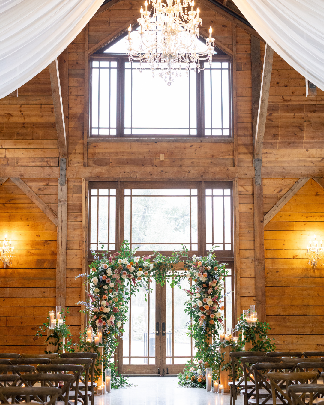 Rustic barn wedding aisle with floral arch, chandeliers, and tall windows under draped white fabric