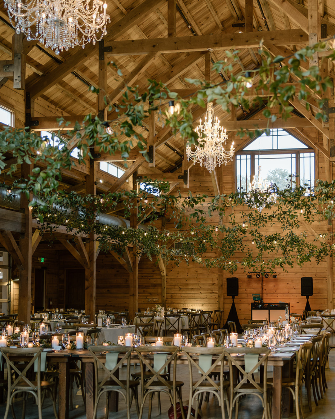 Rustic barn-style wedding reception with long tables, chandeliers, greenery, and warm wooden beams