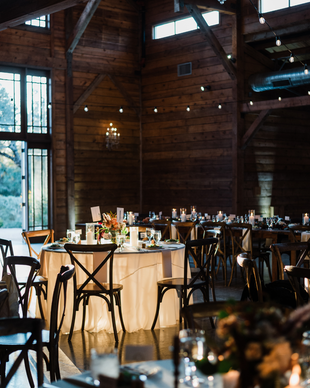 A round table set for a wedding reception in a rustic wooden barn with warm string lighting and dark wooden chairs.