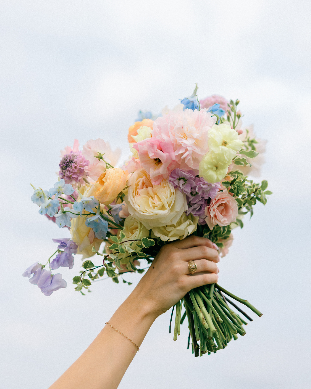 Hand holding a pastel bouquet of mixed flowers against a soft sky background