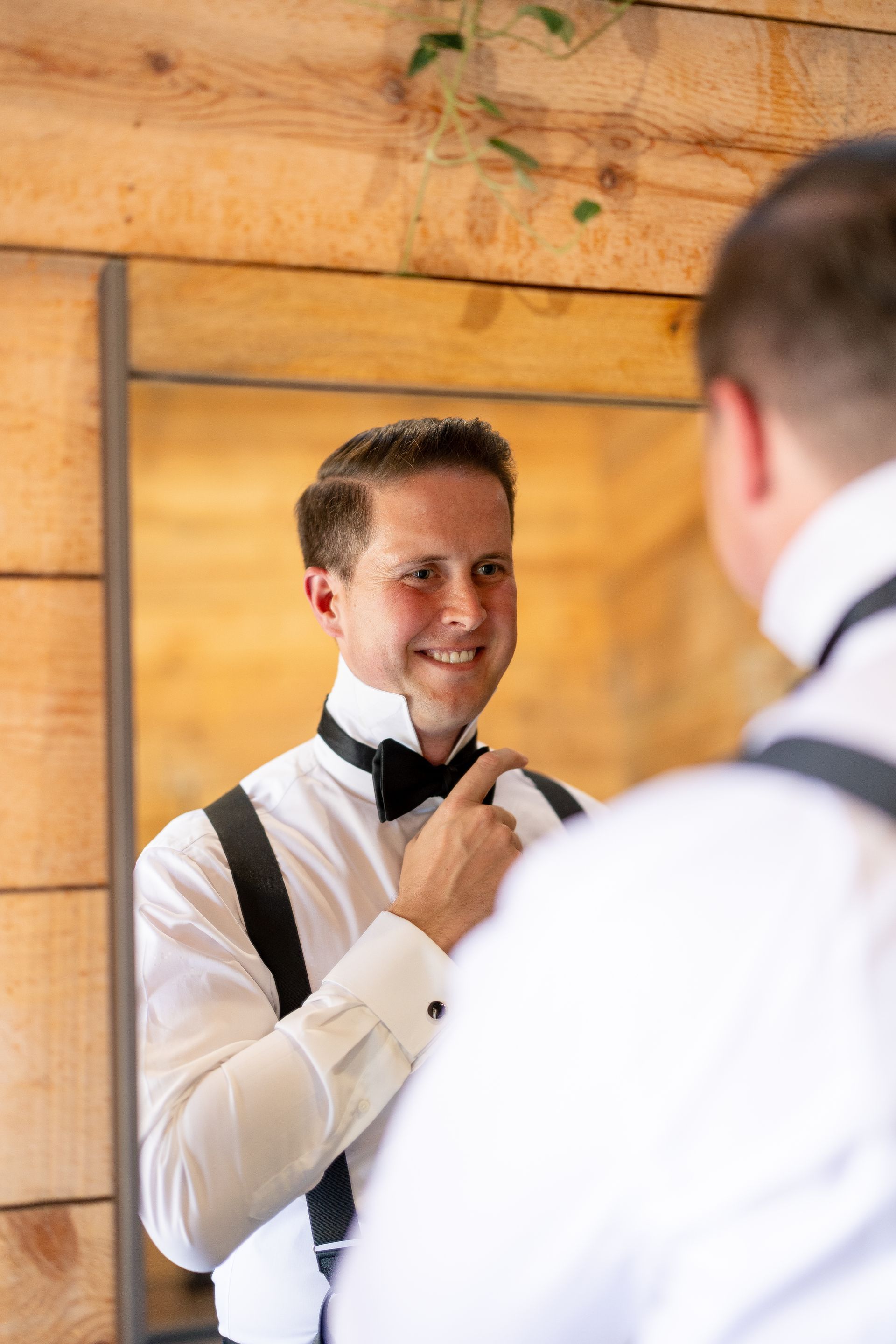 A person in a white dress shirt, black bowtie, and suspenders adjusts their bowtie while looking into a mirror.