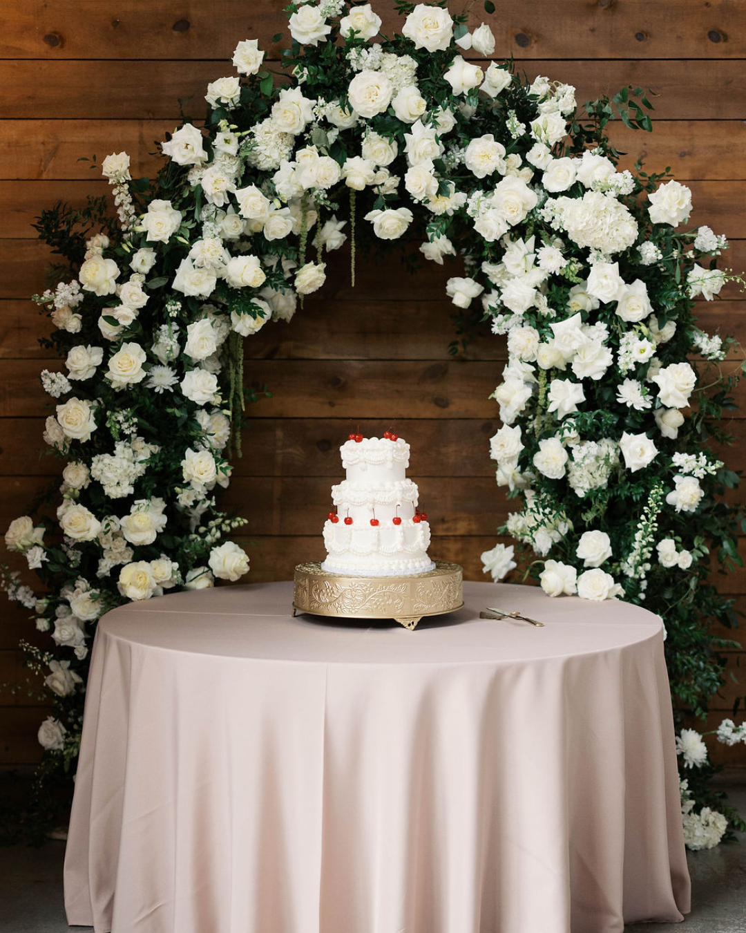 White floral arch behind a small wedding cake on a round draped table against a wooden wall