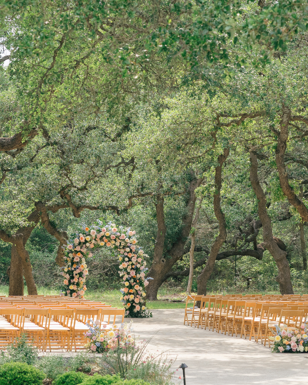 Outdoor wedding setup with floral arch, rows of wooden chairs, and tall trees overhead