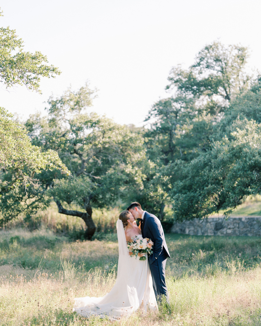 Bride and groom kissing in a sunlit meadow, bride holding a bouquet.