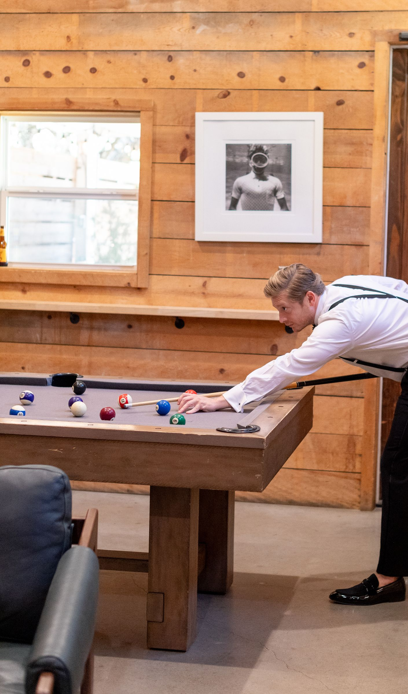 A person in a white button-down shirt and suspenders leans over a wooden pool table to take a shot in a log cabin room.