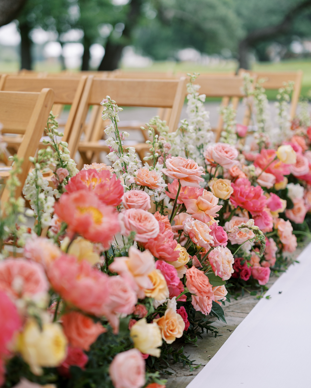 Pink and peach floral arrangement lining an outdoor table with wooden chairs in the background